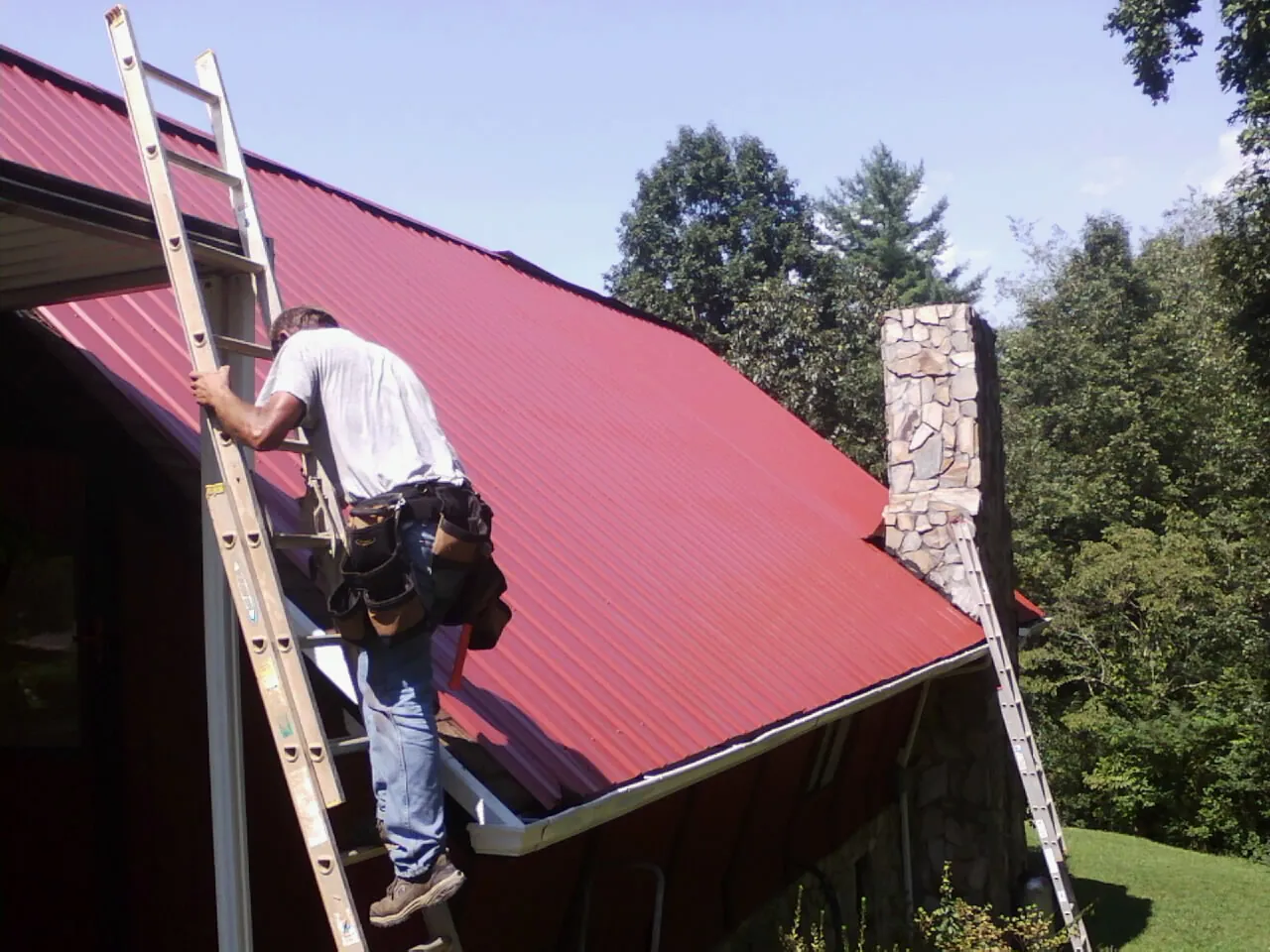 Roofer on a ladder beside a red metal roof and stone chimney — Soffit & Fascia Repair by Roofing Brothers in Liberty Center